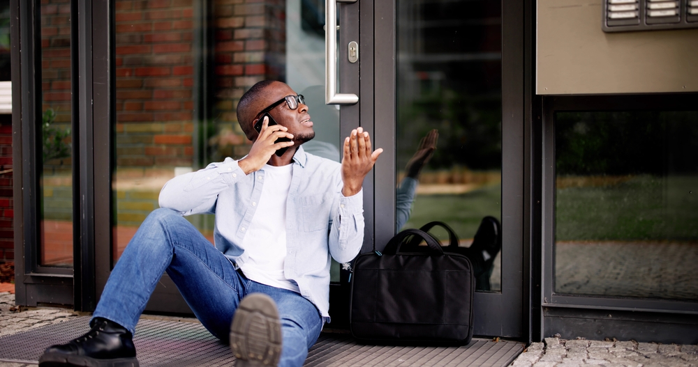 Frustrated man sitting on the ground while on the phone, locked out of his house